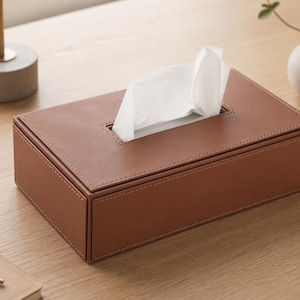 May include: A rectangular brown leather tissue box with white tissues protruding from the top. The box has a stitched border and sits on a light-colored wooden surface. A small white vase with greenery is in the background.
