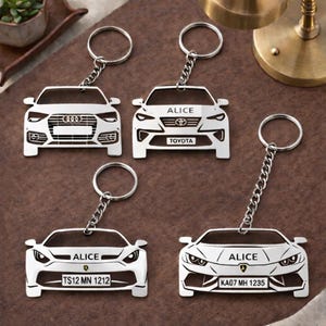 May include: A selection of silver car keyrings. Each keyring showcases a different car model, such as Audi, Toyota, and Lamborghini. Some keyrings are engraved with the name "ALICE" and license plate numbers. The keyrings are displayed on a brown surface.