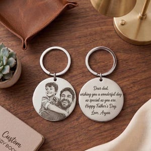 Pode incluir: Dois chaveiros prateados com pingentes redondos. Um chaveiro apresenta uma foto de um homem e uma criança. O outro chaveiro tem o texto "Dear dad, wishing you a wonderful day as special as you are. Happy Father's Day. Love, Aryan."