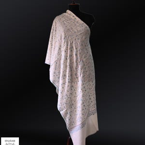 May include: A cream-colored shawl draped over a mannequin, adorned with a delicate blue floral pattern. The shawl features a detailed border design. The brand logo "ITOASH" is visible in the lower left corner. The shawl is made of soft fabric.
