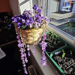Op de afbeelding: Een hangmand gevuld met paarse bloemen en decoratieve kralenstrengen. De mand is gemaakt van geweven materiaal en wordt opgehangen aan een zilveren ketting. Kleine planten staan in groene bakken bij het raam.