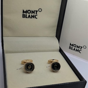 以下が含まれることがあります： 黒い中心部を持つ金色のカフスボタンのペア。"MONTBLANC"のロゴが入った黒い箱に入っています。箱の内側はクリーム色です。カフスボタンは丸く、縁はテクスチャ加工されています。