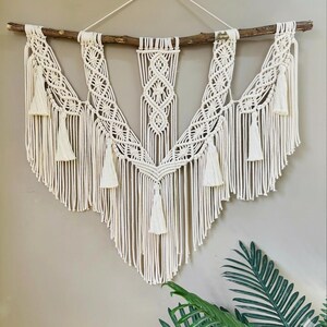 May include: Cream-colored macrame wall hanging with a natural wooden branch. The handcrafted piece features intricate knotting patterns, tassels, and a V-shaped design. This bohemian-style decor adds texture and visual interest to any space.