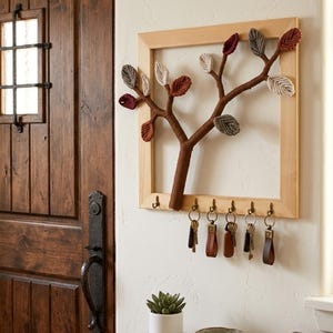Op de afbeelding: Een houten sleutelhouder met een boomontwerp. De boom heeft bruine takken en kleurrijke bladeren in tinten rood, grijs en beige. Onder de boom hangen zes haken met sleutels met leren sleutelhangers. Een kleine vetplant in een witte pot staat eronder.