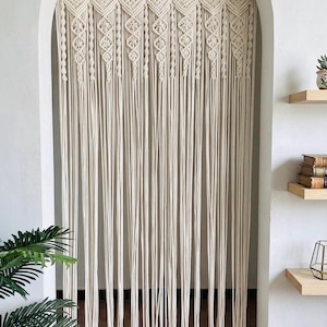 May include: Cream-colored macrame curtain hanging in an arched doorway. The curtain features intricate knotted patterns at the top, with long, vertical strands cascading down. The design adds a bohemian touch to the space. The curtain is made of a natural fiber.