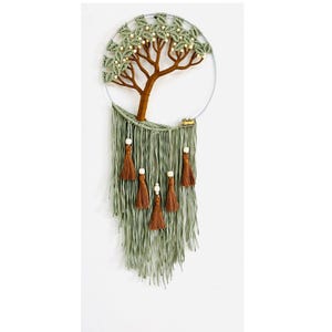 May include: A handmade macramé wall hanging with a tree design. The tree has a brown trunk and green leaves, with white bead accents. Below, long green fringe and brown tassels with white beads hang from a silver hoop.