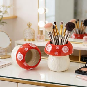 Op de afbeelding: Een rood en wit paddenstoelvormige keramische make-up kwastenhouder en een bijpassend decoratief stuk. De houder bevat make-up kwasten. De paddenstoelkapjes hebben een rode achtergrond met witte stippen.