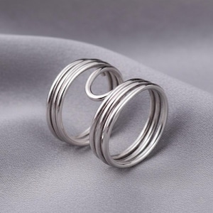 May include: Two silver rings with a unique design. Each ring features multiple parallel bands, creating a textured appearance. One ring has a curved extension that connects to the other ring, adding an artistic touch. The rings are displayed on a gray fabric.