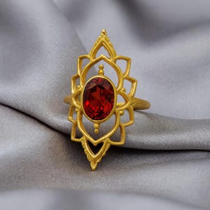 Könnte beinhalten: Ein goldfarbener Ring mit einem ovalen, tiefroten Edelstein in der Mitte. Der Ring hat ein offenes Design mit einem blumen- oder lotusartigen Muster, das den Edelstein umgibt. Der Ring ist vor einem grauen Stoffhintergrund platziert.