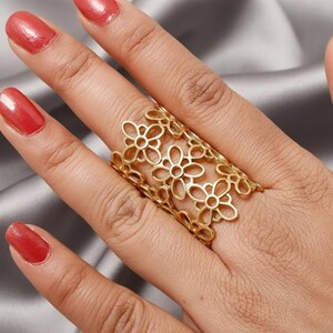 May include: A gold-colored ring featuring a floral design. The ring is worn on a hand with red nail polish. The ring's design consists of multiple openwork flower shapes, creating a decorative and airy appearance. The ring is a statement piece.