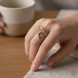 May include: A gold-colored ring with a heart-shaped design. The ring is worn on a finger and features a stylized letter "A" with a heart incorporated into the design. The ring has a textured surface and is set against a neutral background.