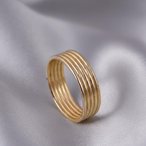 Puede incluir: Un anillo dorado con un diseño multibanda. El anillo presenta cinco bandas finas y paralelas, creando una superficie texturizada. El anillo está sobre un fondo suave gris plateado.