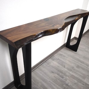 Puede incluir: Una mesa consola con un tablero largo de madera marrón oscuro con un borde natural y un acabado liso y pulido. La mesa se apoya sobre dos patas de metal negro, geométricas y de marco abierto, que proporcionan una base estable a la superficie.