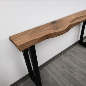 Pode incluir: Uma mesa consola longa com um tampo espesso de madeira natural de borda viva, com padrões de grão visíveis e acabamento castanho escuro. A mesa é suportada por duas pernas de metal preto, cada uma desenhada com uma estrutura geométrica angular.