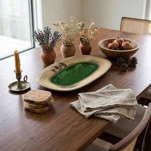 Pode incluir: Uma mesa de jantar de madeira com um castiçal de latão com uma vela amarela, três pequenos vasos de terracota com flores secas, uma bandeja oval de madeira com centro esmaltado verde, uma tigela de madeira com romãs e guardanapos de linho.