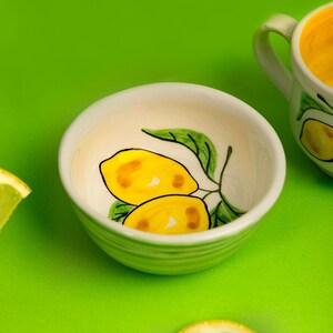 Może przedstawiać: Mała, biała ceramiczna miska z ręcznie malowanym wzorem cytryny. Miska przedstawia dwie żółte cytryny z zielonymi liśćmi. Plasterek cytryny znajduje się na pierwszym planie, a pasujący kubek w tle, wszystko na zielonej powierzchni.