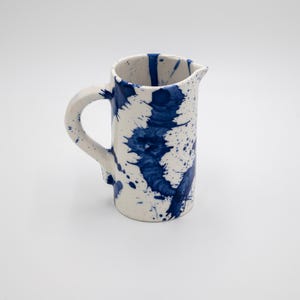 Peut inclure: Un pichet en céramique avec une base blanche et un motif d'éclaboussures de peinture bleu foncé. Le récipient possède une anse incurvée sur le côté et un petit bec verseur sur le bord supérieur. Le motif bleu recouvre l'extérieur et l'intérieur.