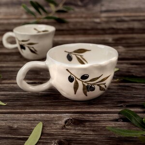 Puede incluir: Dos tazas de cerámica color blanco roto con una forma irregular hecha a mano. Cada taza presenta un diseño pintado de aceitunas negras y hojas de olivo verdes tanto en el exterior como en el interior. Tienen una asa redondeada en el lateral.