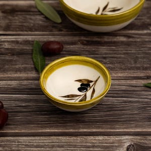 Puede incluir: Dos cuencos redondos de cerámica con interior blanco y borde pintado en verde. Cada cuenco presenta un diseño pintado a mano de una rama de olivo marrón con dos aceitunas negras en el interior. Los cuencos están sobre una superficie de madera.