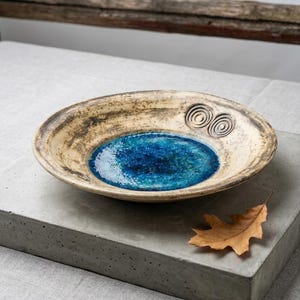 Pode incluir: Uma tigela de cerâmica rasa com uma borda texturizada de cor creme apresentando dois padrões de espiral escuros. O centro da tigela tem um esmalte azul profundo e brilhante com uma aparência rachada. Forma e acabamento orgânicos.