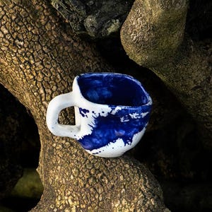 Op de afbeelding: Een witte keramische mok met een dik, afgerond handvat en een onregelmatige, organische vorm. De binnen- en buitenkant zijn voorzien van een levendig, diepblauw glazuur in een spetterpatroon dat contrasteert met de witte basiskleur.