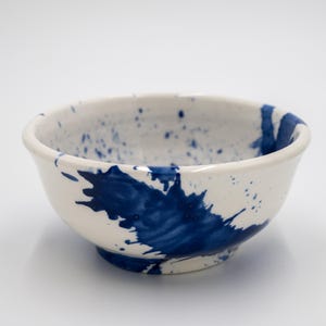 May include: A white ceramic bowl featuring a glossy finish and a bold, dark blue paint splatter pattern. The design includes large, irregular blue splashes on the exterior and interior surfaces, creating a high-contrast, artistic appearance.