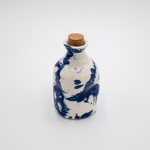 Könnte beinhalten: Eine kleine Keramikflasche mit weißem Untergrund und einem dunkelblauen Farbspritzmuster. Die Flasche hat einen abgerundeten Körper, einen schmalen Hals und einen zylindrischen Korkstopfen, der in die obere Öffnung eingesetzt ist.
