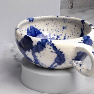 Op de afbeelding: Een keramische mok met een glanzende gebroken witte basis en een levendig blauw verfspattenpatroon. De mok heeft een brede, ronde komvorm en een enkel gebogen handvat aan de rechterkant, waarbij de blauwe spatten zowel binnen als buiten zitten.