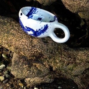 Op de afbeelding: Een witte keramische mok met een levendig blauw spetterpatroon op het buiten- en binnenoppervlak. De mok heeft een afgerond, dikwandig lichaam en een stevig, cirkelvormig handvat aan de zijkant, afgewerkt met een glanzende glazuurlaag.