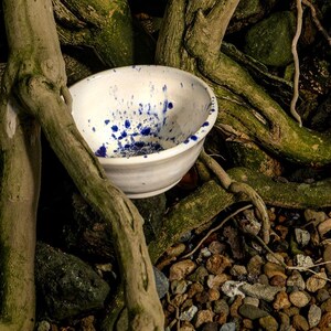Op de afbeelding: Een witte keramische kom met een gespikkeld blauw verfspatpatroon op het binnenoppervlak. De kom heeft een gladde, afgeronde rand en een licht taps toelopende voet, wat zorgt voor een strak en functioneel ontwerp met kleurcontrast.