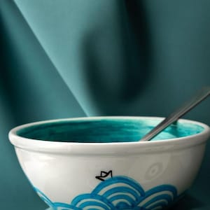 Puede incluir: Un cuenco de cerámica blanca con interior verde azulado y un patrón de olas azules pintado a mano en el exterior. Un pequeño dibujo lineal negro de un barco de papel se sitúa sobre las olas. Una cuchara de metal plateado descansa en el cuenco.