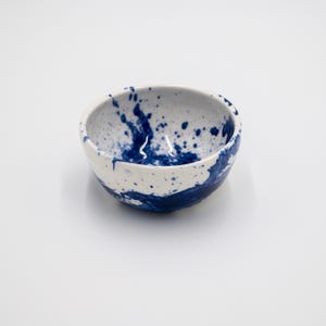 Op de afbeelding: Een kleine, ronde keramische kom met een witte basis en een levendig blauw verfspattenpatroon. De binnen- en buitenoppervlakken zijn versierd met onregelmatige blauwe stippen en grotere spatten van blauw pigment over het witte glazuur.