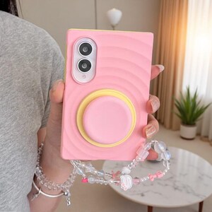 May include: A rectangular pink phone case with a wavy texture, a yellow circular pop-out grip, and a dual-lens camera cutout. A beaded wrist strap with clear, white, and pink beads, including a small pink bow and a charm, is attached to the case.