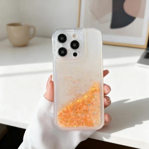 May include: Clear phone case with a liquid glitter design in shades of orange and gold. The case has a transparent border and a camera cutout. The case is held in a hand.