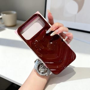 May include: A burgundy phone case featuring a heart design. The case has a glossy finish and a raised heart pattern. The case is held against a pink and white background.