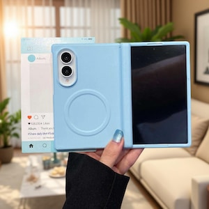 Puede incluir: Una funda de smartphone plegable de color azul claro con un anillo magnético circular en la parte posterior y un recorte para cámara de doble lente. La funda se muestra junto a una tarjeta con una interfaz de red social con el texto Album, 520,1314 Likes y #Share daily life.