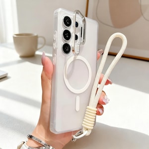 May include: Clear phone case with a silver metal ring and a white lanyard. The case has a white circle and a vertical line design. The lanyard is made of a white rope with a cream-colored, ribbed accent.