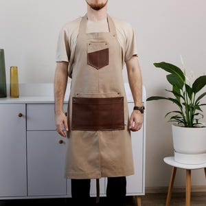 Pode incluir: Avental bege com bolsos de couro castanho. O avental tem um bolso retangular no peito e um bolso grande retangular na cintura. O avental é usado sobre uma camiseta bege e calças pretas. A pessoa está usando um relógio e tênis.