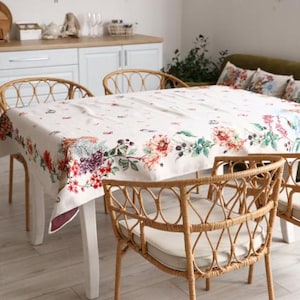 Puede incluir: Un comedor con una mesa cubierta con un mantel blanco adornado con un estampado floral en tonos rojos, morados y verdes. Cuatro sillas de mimbre rodean la mesa. La habitación tiene un ambiente luminoso y espacioso.