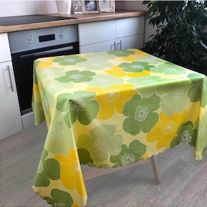 May include: A square tablecloth with a floral pattern in shades of yellow and green. The flowers have a white center and are scattered across the fabric. The tablecloth is draped over a wooden table.