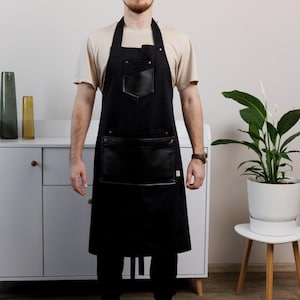 Może przedstawiać: Czarny fartuch z skórzanymi kieszeniami. Fartuch ma pasek na szyję i wiązania w talii. Model ma na sobie beżowy t-shirt, czarne spodnie i trampki. Fartuch jest przeznaczony do użytku w kuchni lub warsztacie.