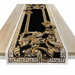 Puede incluir: Camino de mesa rectangular negro con diseños florales barrocos dorados y un borde con patrón de cadena. El camino de mesa tiene un borde blanco con un patrón de llave griega marrón. Sobre una mesa de madera clara.