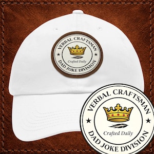 Op de afbeelding: Witte baseballpet met een bruine patch. De patch heeft de tekst "VERBAL CRAFTSMAN DAD JOKE DIVISION", een gouden kroon, de woorden "Crafted Daily" en sterren.