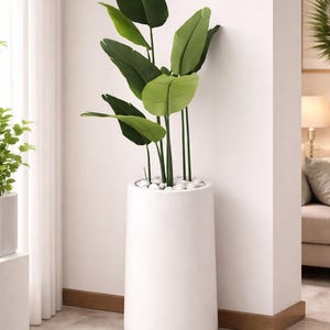 May include: A large, white cylindrical planter contains an artificial plant with broad, green leaves. The plant's stems are dark green, and white pebbles cover the soil. The planter is set against a white wall, creating a minimalist aesthetic.