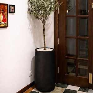 May include: A tall, black cylindrical planter holds an artificial olive tree. The tree has a slender brown trunk and light green leaves. The planter sits on a wooden floor next to a white wall and a dark wooden door.