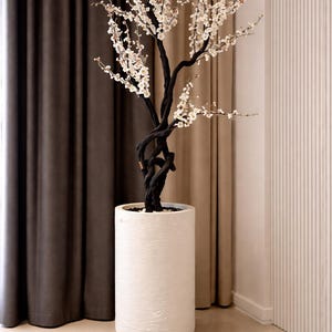 Puede incluir: Un árbol artificial decorativo con flores blancas y un tronco oscuro y retorcido. Está plantado en una maceta cilíndrica alta, blanca y texturizada. El fondo incluye cortinas marrones y beige y una pared blanca.