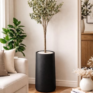 May include: A tall, artificial tree in a black cylindrical planter. The tree has a slender trunk and green and white leaves. The planter is filled with small white stones. The tree is in a well-lit room with a sofa and a round coffee table.
