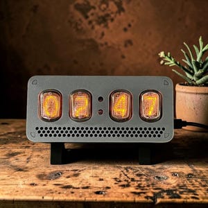 Könnte beinhalten: Eine graue Digitaluhr mit vier leuchtenden orangefarbenen Röhren, die Zahlen anzeigen. Die Uhr hat ein rechteckiges Zifferblatt mit perforiertem Boden und steht auf einem schwarzen Ständer. Eine kleine Topfpflanze befindet sich im Hintergrund.