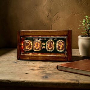 Könnte beinhalten: Eine digitale Holzuhr, die "20:09" mit leuchtenden orangefarbenen Ziffern anzeigt. Die Uhr, mit der Aufschrift "GeekStyles IN12 U3", steht auf einer Holzoberfläche, mit einer kleinen Topfpflanze und einem Buch im Hintergrund. Die Uhr hat eine Retro-Ästhetik.