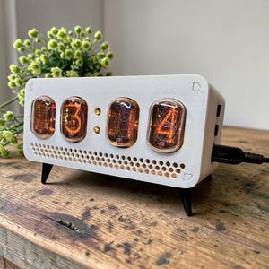 Puede incluir: Un reloj digital blanco con cuatro tubos luminosos de color naranja que muestran la hora. El reloj tiene una parte delantera perforada y se apoya en pequeñas patas negras. Un cable USB negro está enchufado en el lateral. El reloj está sobre una superficie de madera.